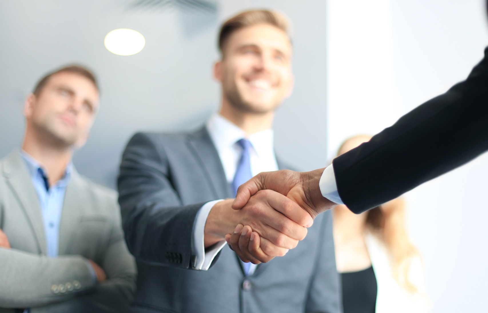 Two business professionals shake hands in a bright office. Colleagues observe, indicating a successful meeting or partnership negotiation.