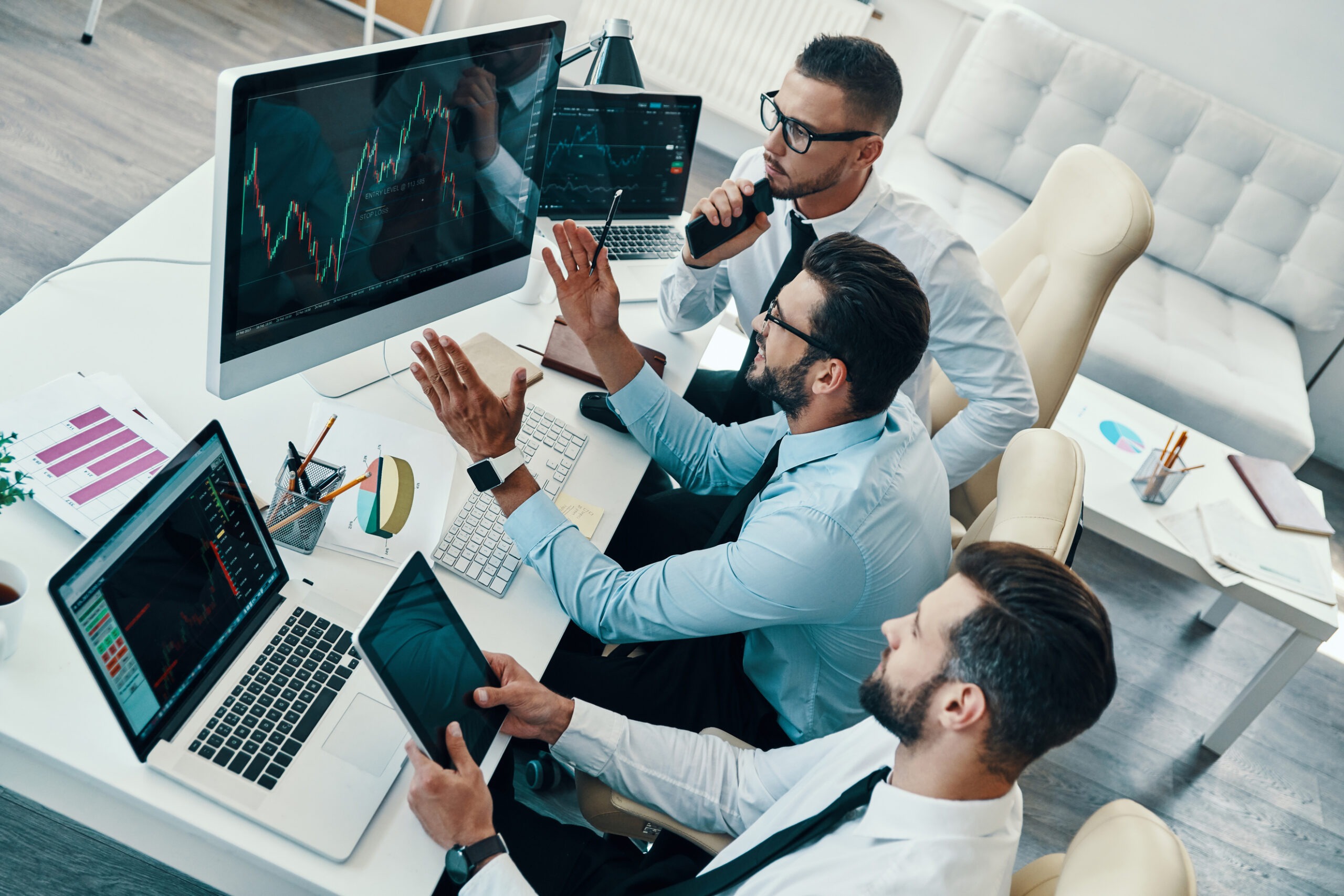 Three business professionals analyze stock charts on multiple screens, discussing strategies in a modern office setting.