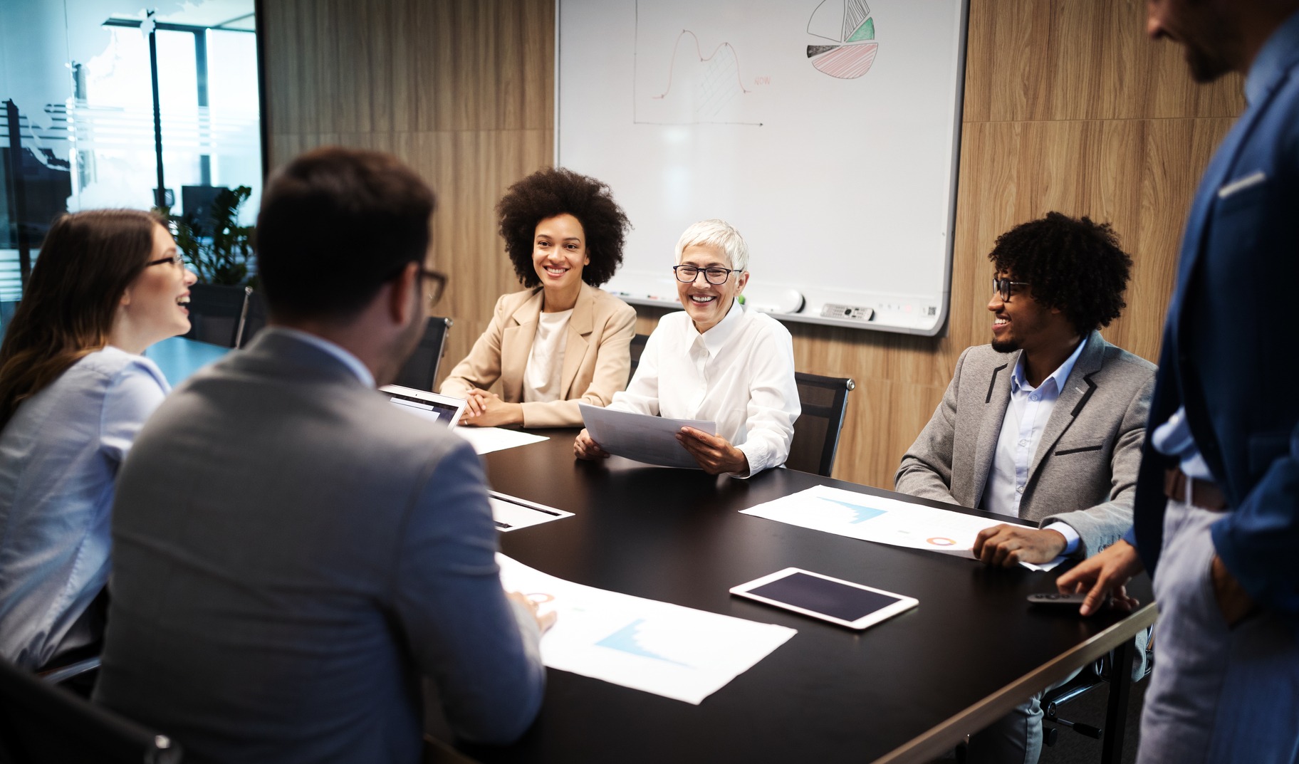 A diverse group of professionals engaged in a meeting, discussing documents and data analysis, fostering collaboration in a modern office setting.
