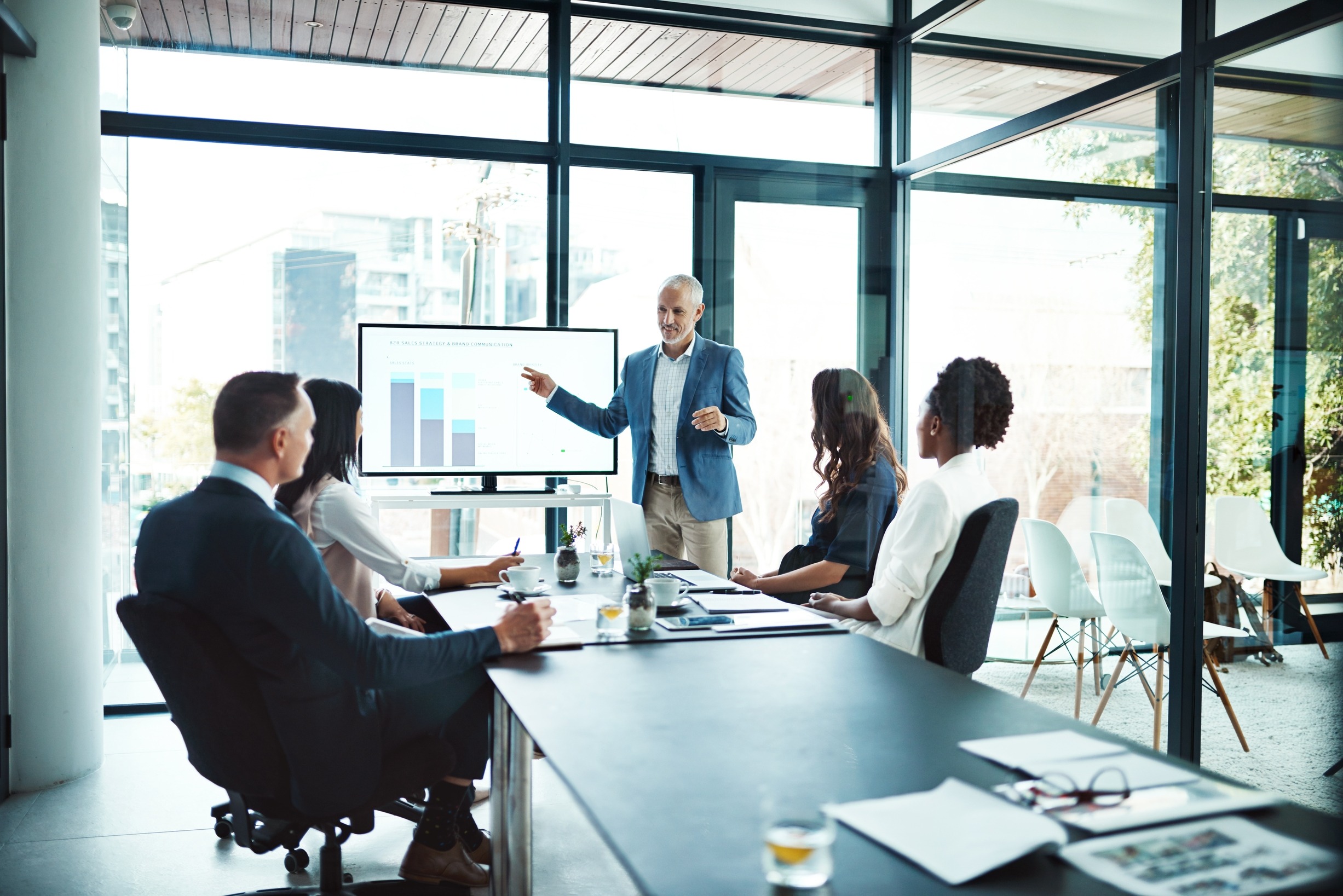 A business presentation in a modern conference room. A speaker presents data on a screen to attentive colleagues seated around a table.
