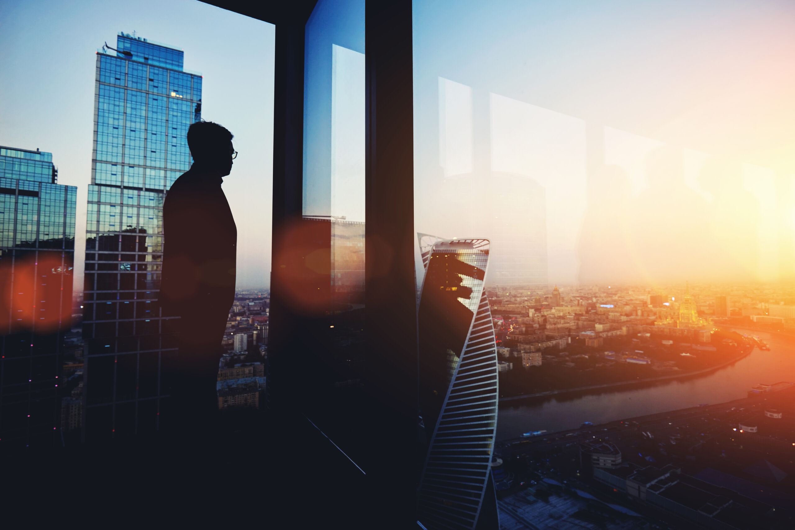 Silhouette of a person gazing out a tall building window at a cityscape during sunset, highlighting modern architecture and reflective glass.