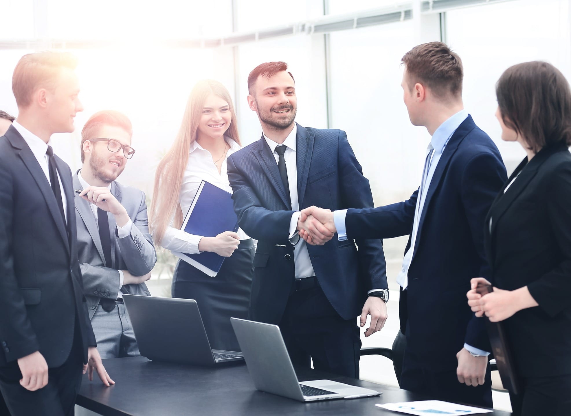 Business professionals engaged in a handshake, symbolizing a partnership or agreement, with laptops and colleagues in the background.