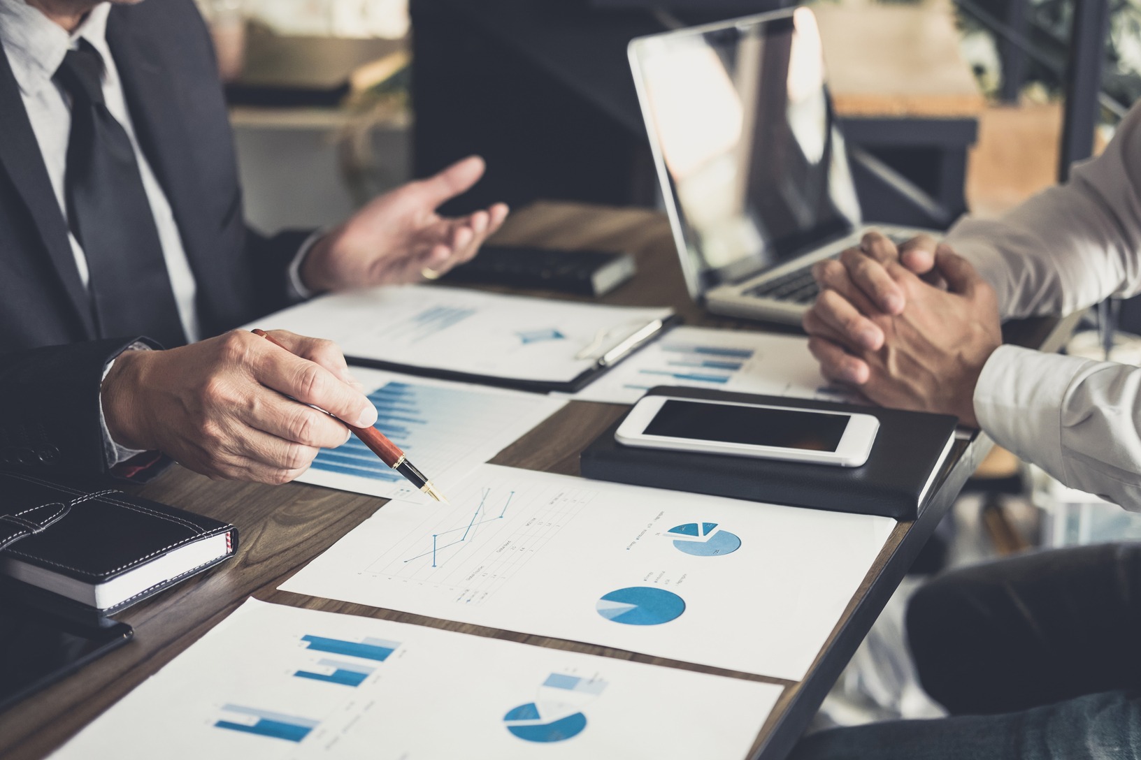 Business professionals discussing financial graphs and data during a meeting, with a laptop and mobile device on the table.